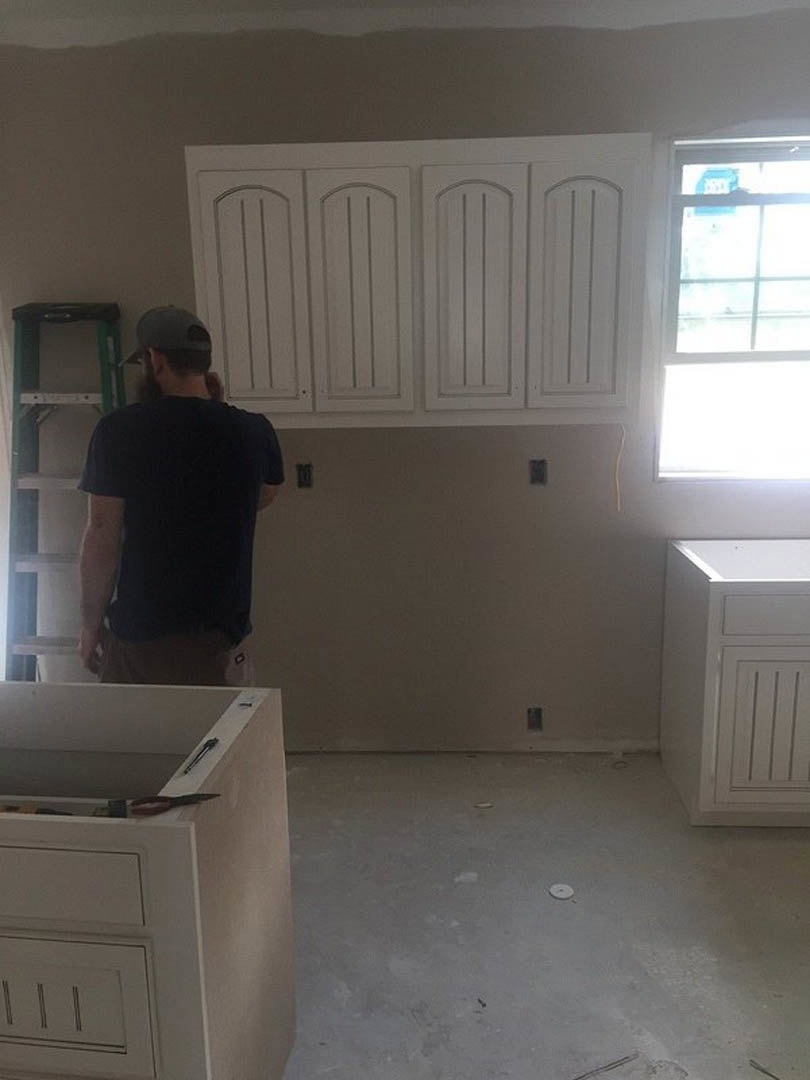 Man in black shirt standing in modern kitchen with white cabinets, vent hood, window with white frame, and countertop holding a knife and pencil.