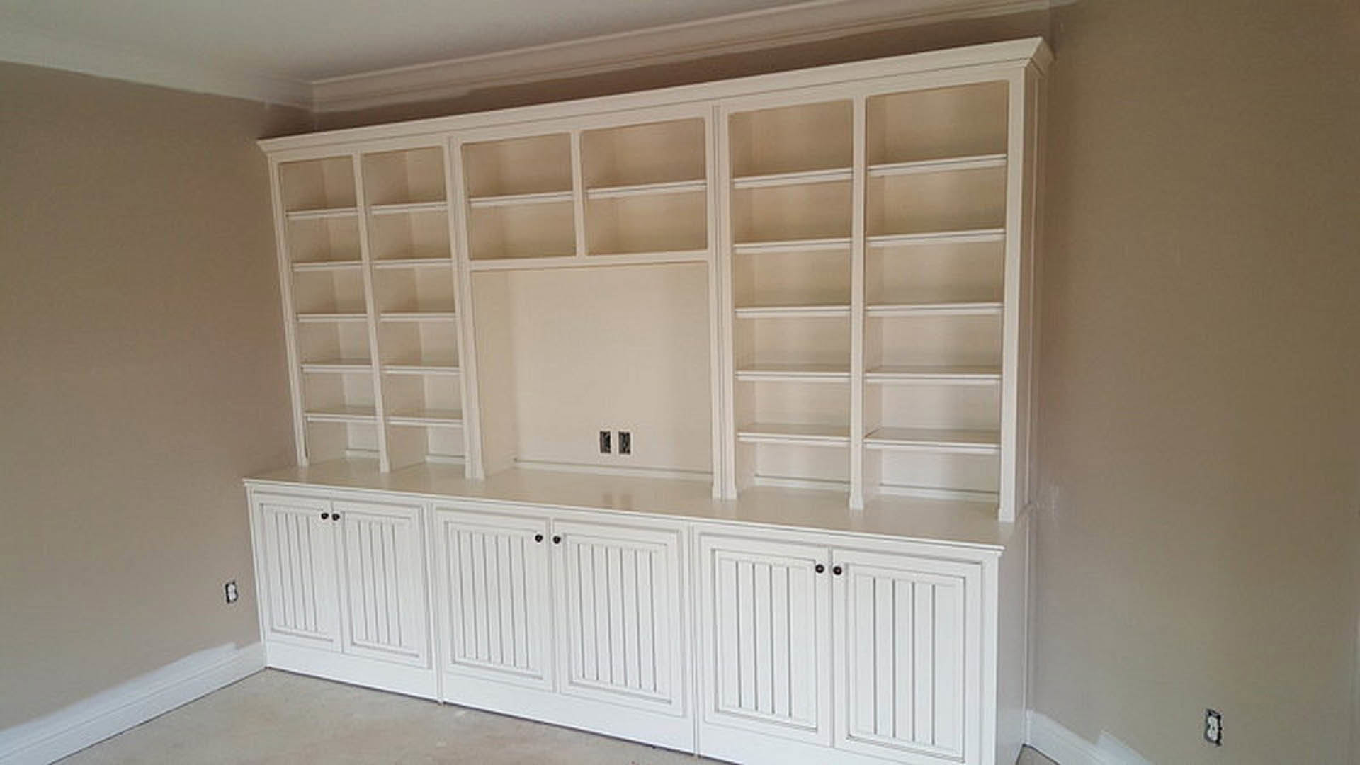 White built-in shelves and cabinet with black knobs against a white wall, hardwood flooring, window, and white electrical outlet.