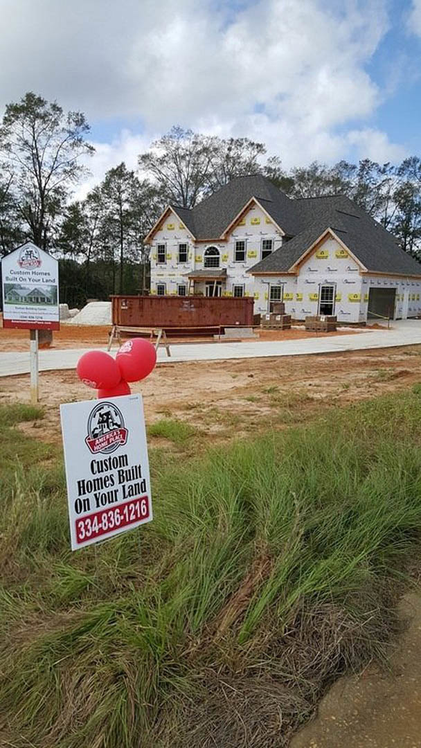 Partially built house with exposed framing, red brick wall, yellow construction signs, white text, balloons, and grassy lawn under cloudy sky