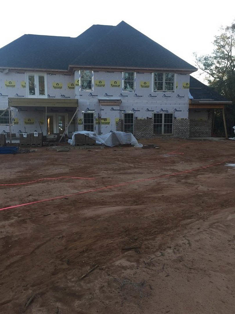 Framed house under construction with red tape marking entry, yellow caution signs on exterior, dirt ground with worker, white bag on table, partially finished roof, windows, and