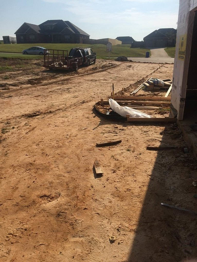 Partially constructed house with exposed roof framing, construction truck and trailer parked on dirt lot, scattered building materials, grassy area and trees in background