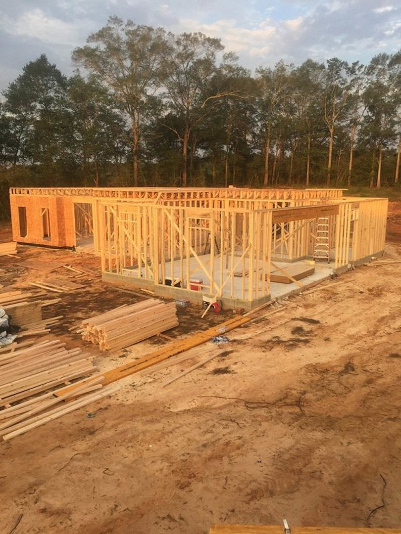 Wood-framed house under construction with exposed beams, stacks of lumber on dirt foundation, surrounded by tall trees and cloudy sky