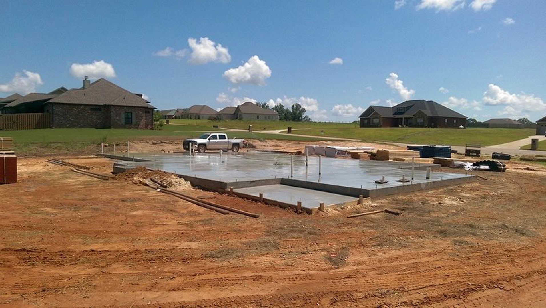 Silver pickup truck parked beside a partially constructed house with gray roof, red exterior wall, and brick chimney; grassy lot with scattered construction materials, trees, and
