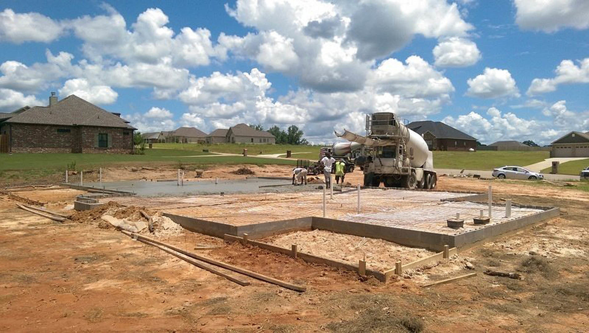 Partially built house with brick chimney, cement truck parked on dirt lot, workers in green shirts walking near construction materials, cloudy sky overhead, trees and grass