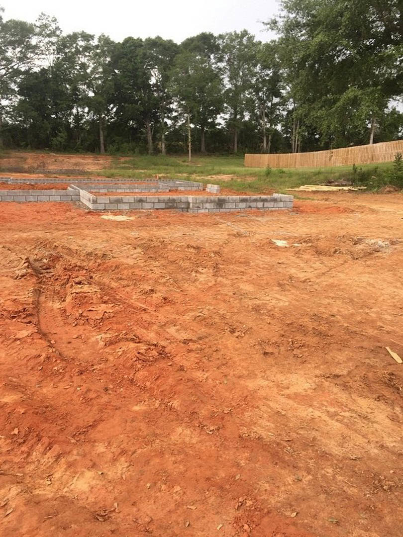 Dirt lot with scattered bricks, temporary fence, and mature trees bordering the construction site