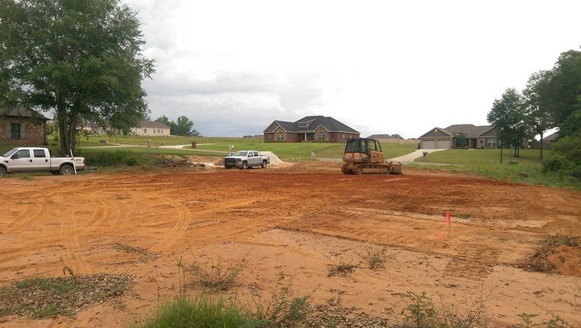 Bulldozer grading dirt near a partially constructed house with black roof, white truck parked nearby, trees and grass in background