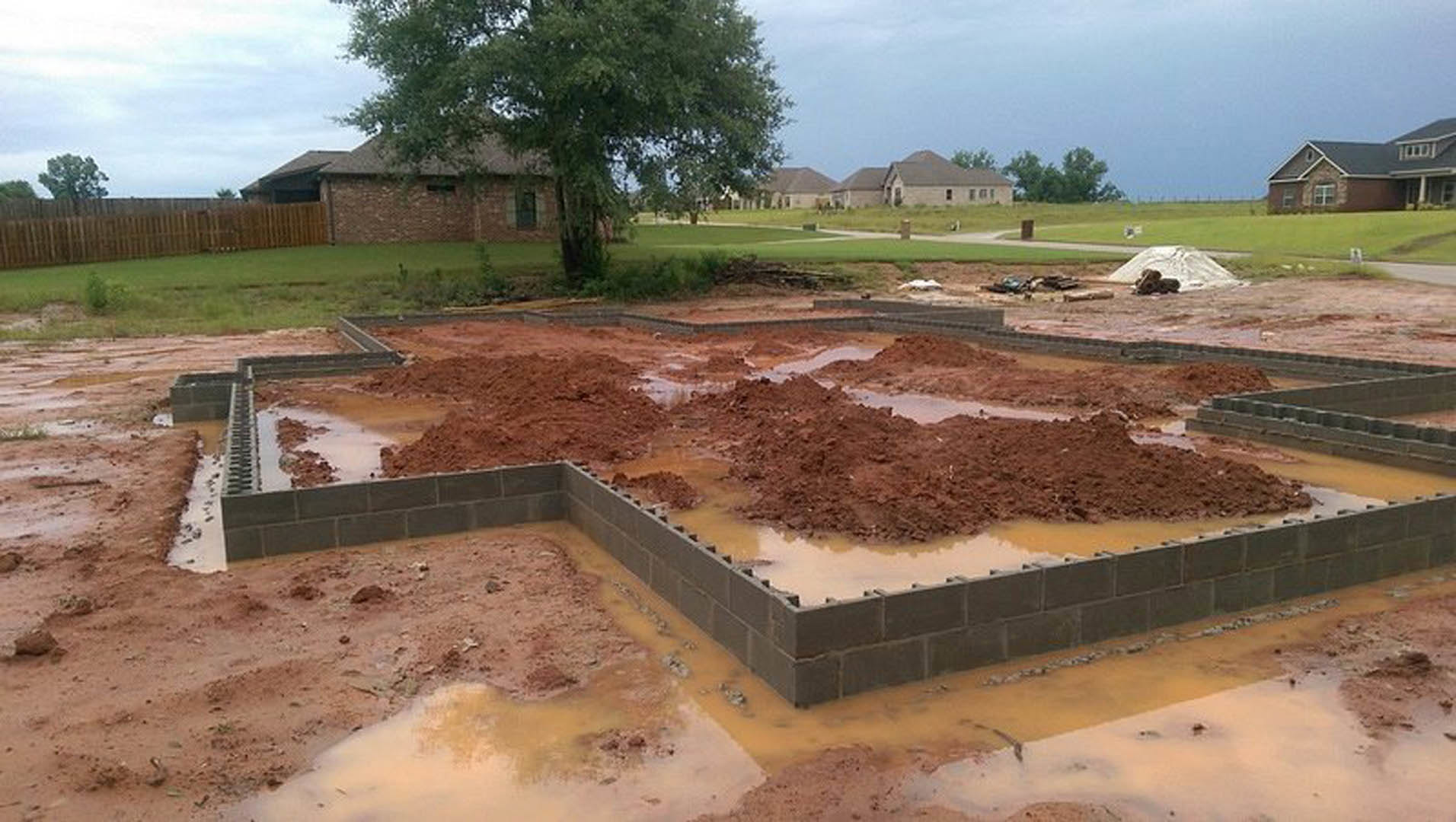 Concrete house foundation surrounded by muddy soil, construction site with scattered water, tree in front of partially built brick structure, wooden fence in background, blue sky