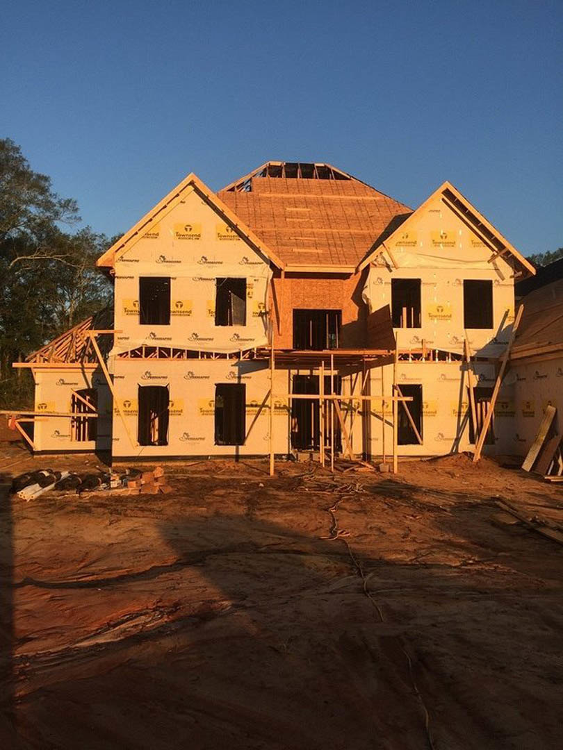 Wood-framed house under construction with scaffolding, exposed roof beams, dirt ground, wooden posts, and several people working near a tree and temporary fencing