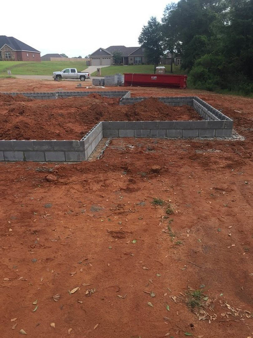 Brick house with black roof under construction, white truck parked on dirt ground, concrete foundation visible, scattered bricks and construction materials, trees and sky in