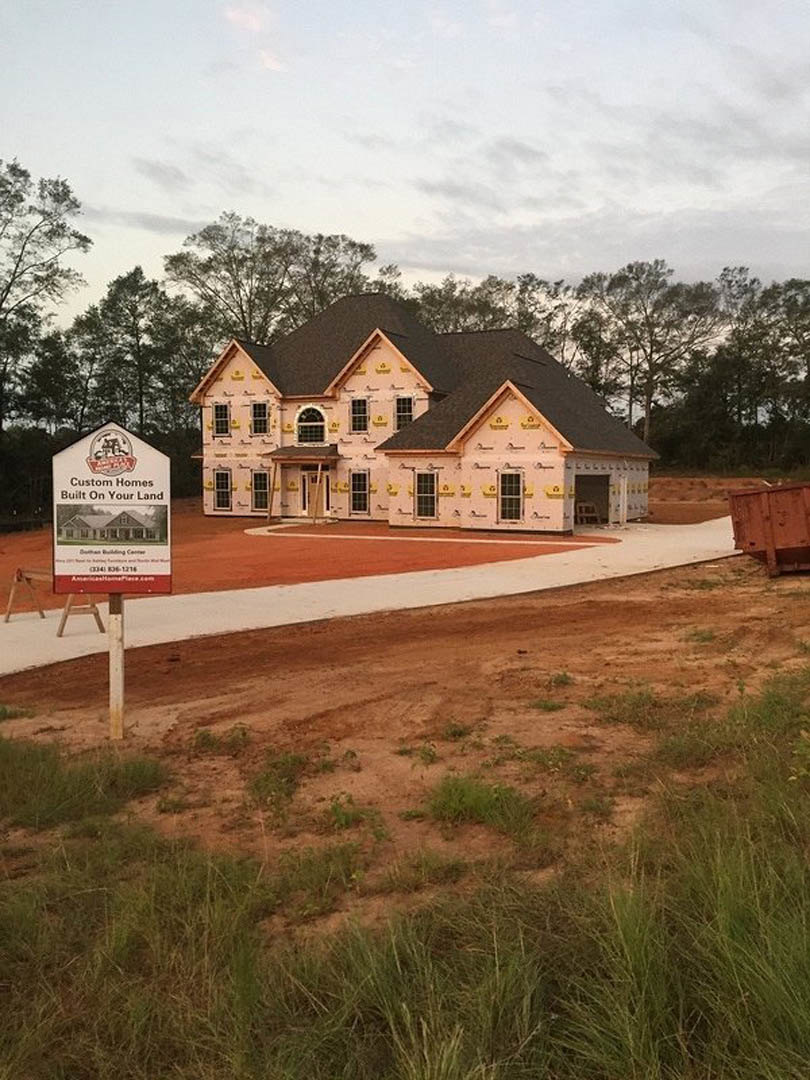 Two-story house under construction with black roof, exposed wooden framing, and a sign on a stand in a dirt lot with patches of grass and trees in the background