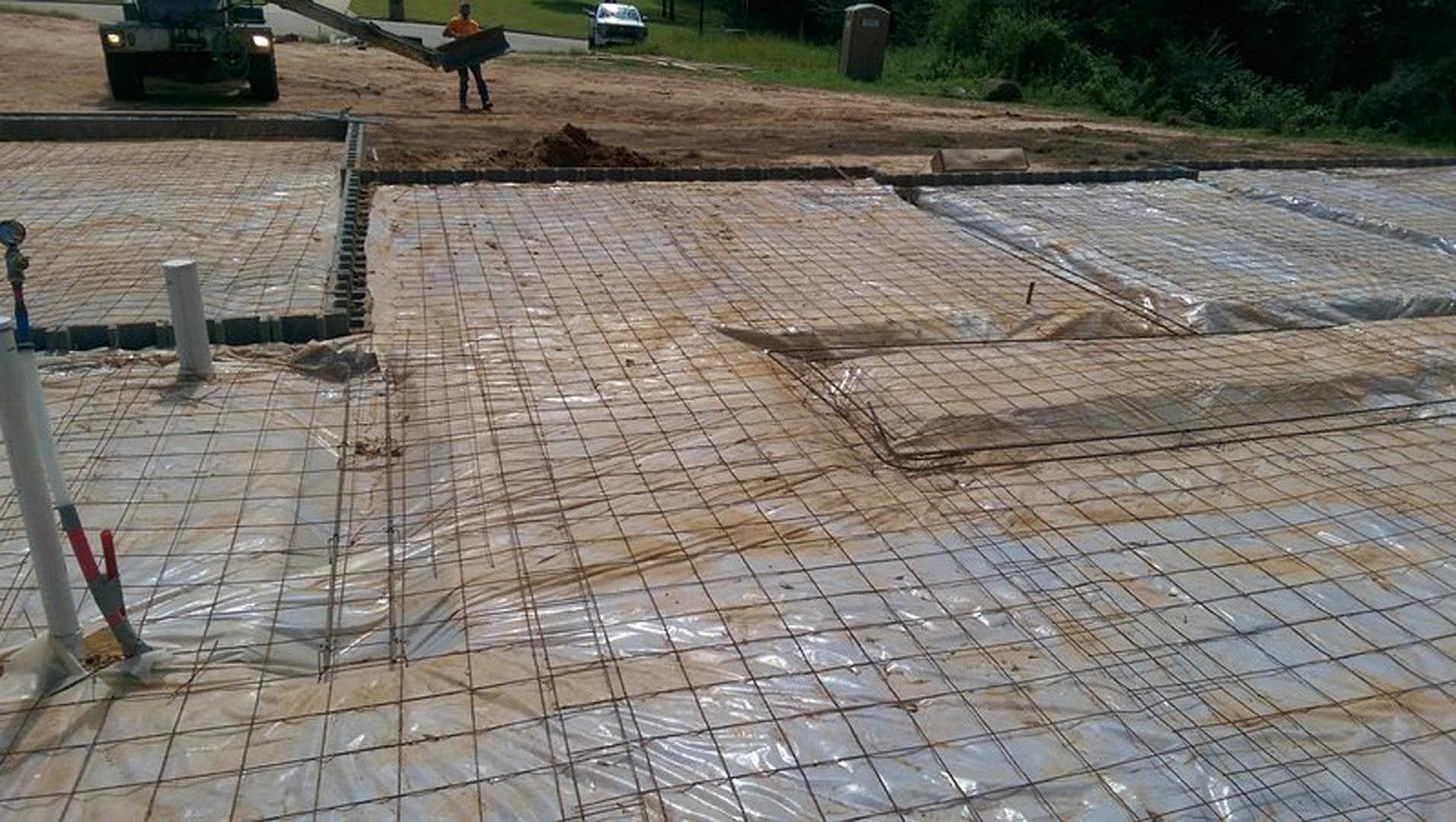 Multiple men standing together on a construction site with concrete, flagstone, and composite materials visible on the ground; trees and building structure in the background.