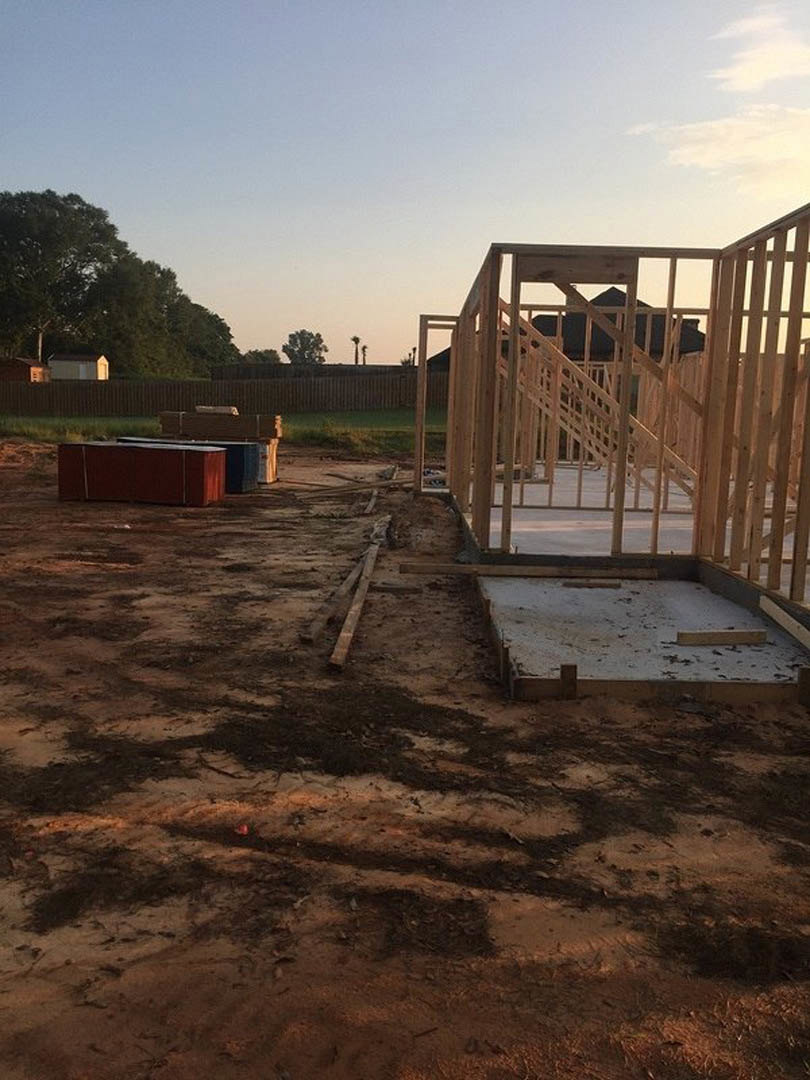 Wood-framed custom home under construction with exposed beams, concrete slab foundation, partially built stairs, dirt ground, and trees visible in the background.