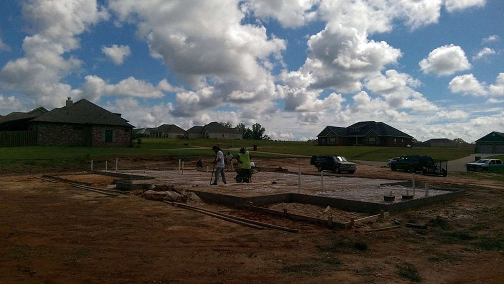 Framed custom home under construction with workers on site, black van and car parked nearby, grassy lot, cloudy sky, and distant mountains in rural landscape