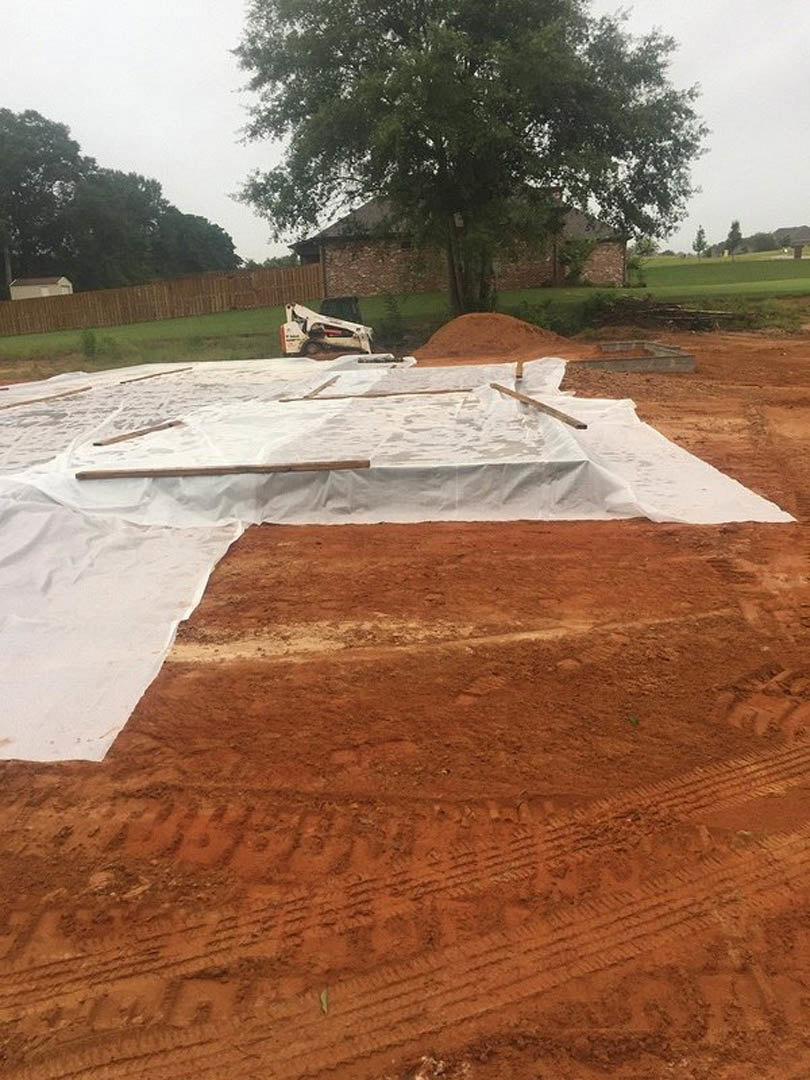 White tarp draped over construction materials beside a dirt road, tree in front of partially built house, white truck parked near green field and wooden fence