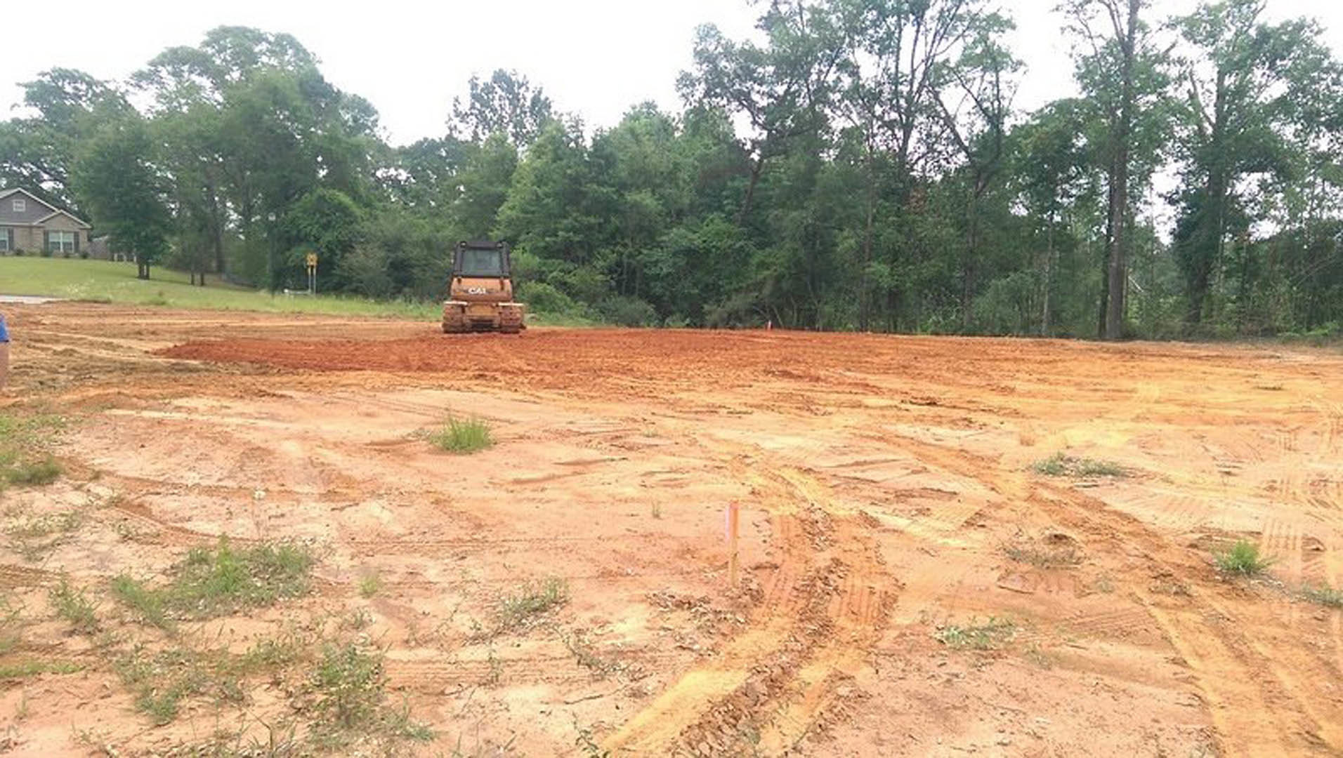 Bulldozer parked on a muddy dirt lot with scattered trees and a partially visible custom home in the background