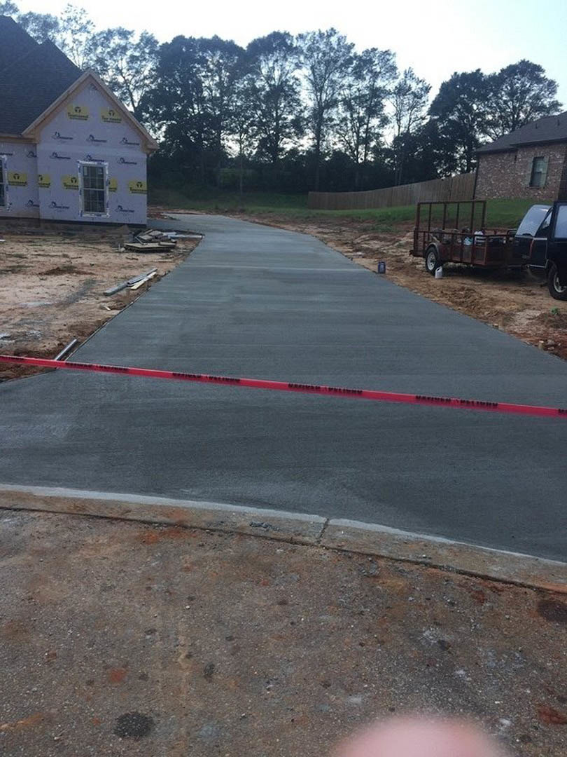 Asphalt road with red caution tape in foreground, partially constructed custom home with yellow and black logo signage, truck and trailer parked nearby, trees and sky in background