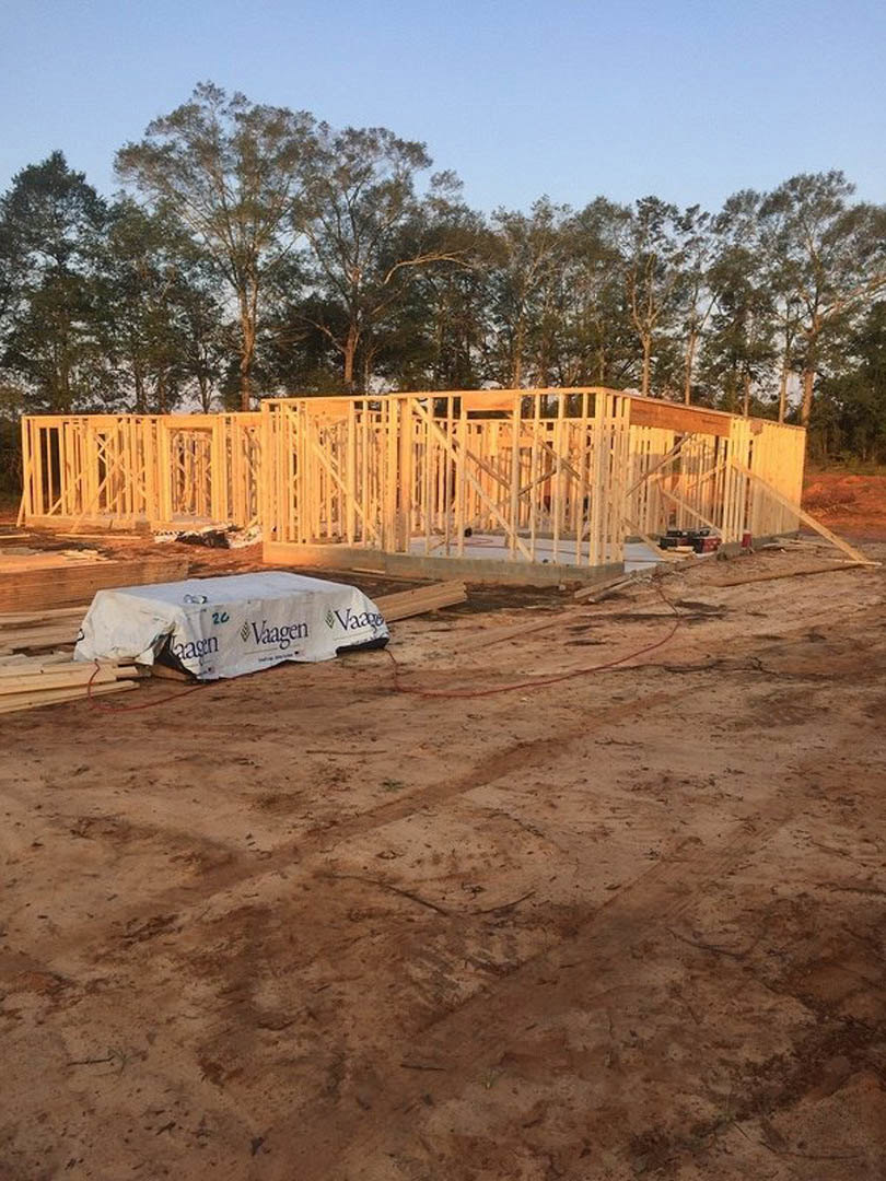 Wood-framed house under construction with exposed beams, large white tarp covering dirt foundation, surrounding trees and clear blue sky in background