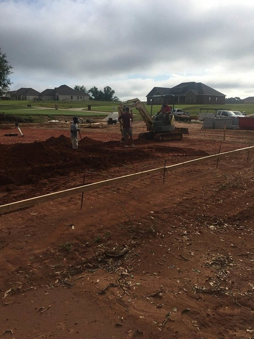 Bulldozer parked on dirt construction site beside partially built house with shingled roof, fenced perimeter, scattered construction materials, and cloudy sky overhead.