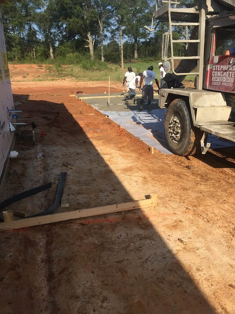 Group of people gathered on a dirt road near a parked truck, surrounded by trees and soil, with visible tire tread and metal rim, one person wearing a white shirt