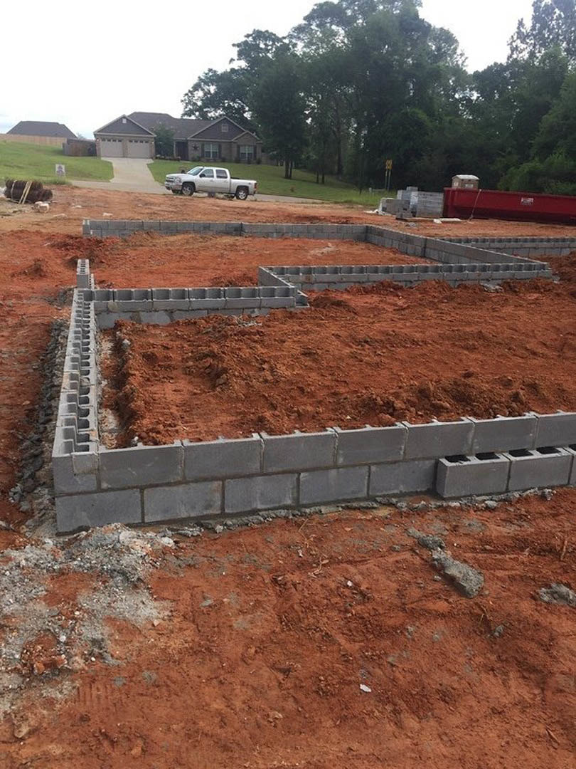 Concrete foundation surrounded by red dirt, white truck parked nearby, partially built house with garage, trees in background, red container on dirt road