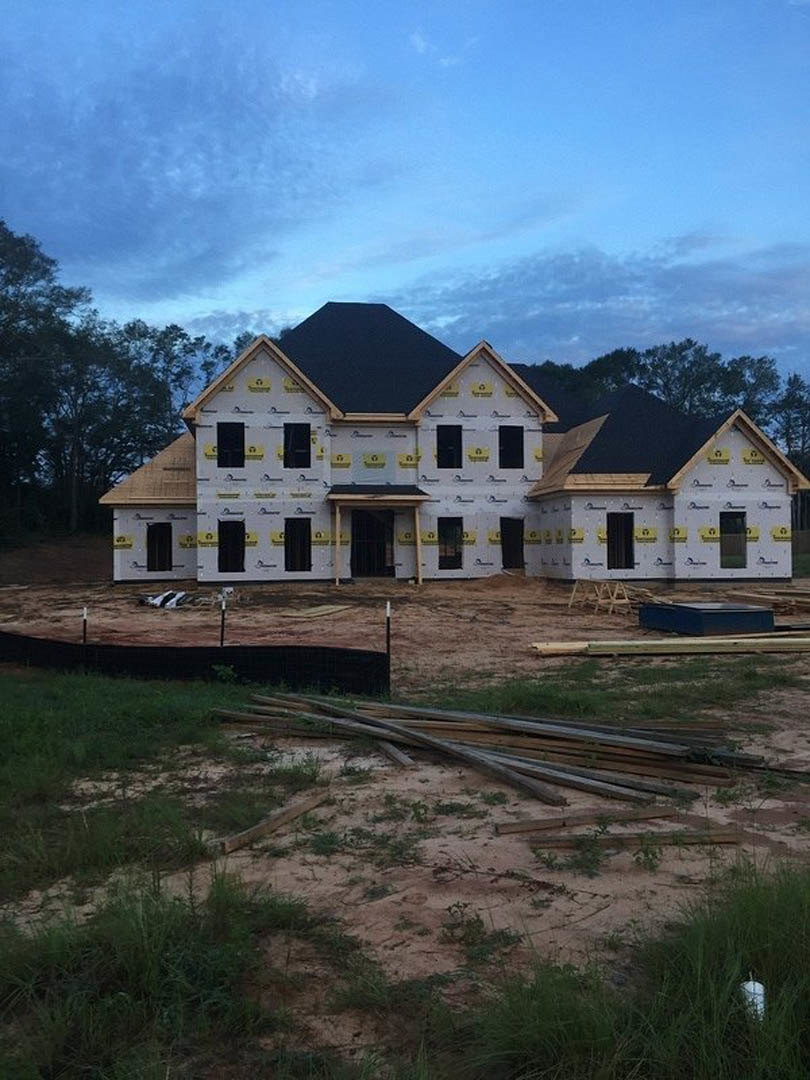 Framed house under construction with black roof, surrounded by trees and grass, pile of lumber in foreground, blue sky with scattered clouds
