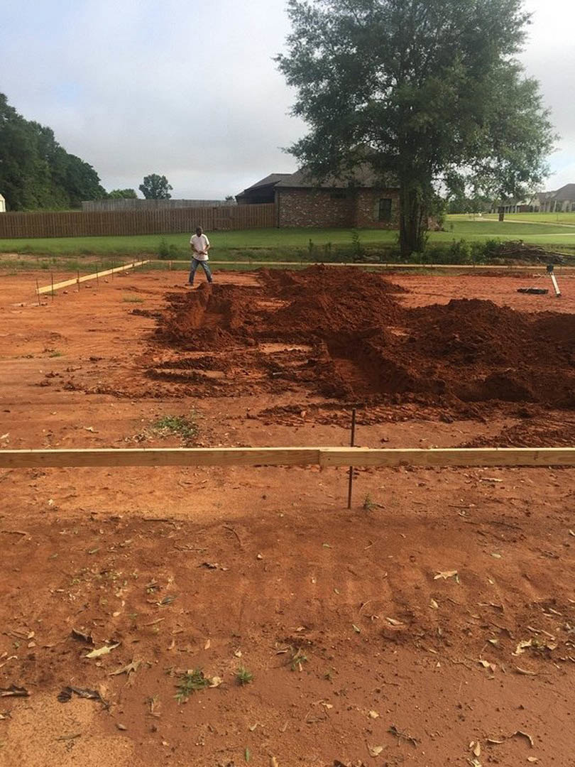 Brick house with tree in front, dirt yard bordered by wooden fence, patches of grass and pile of soil in open field