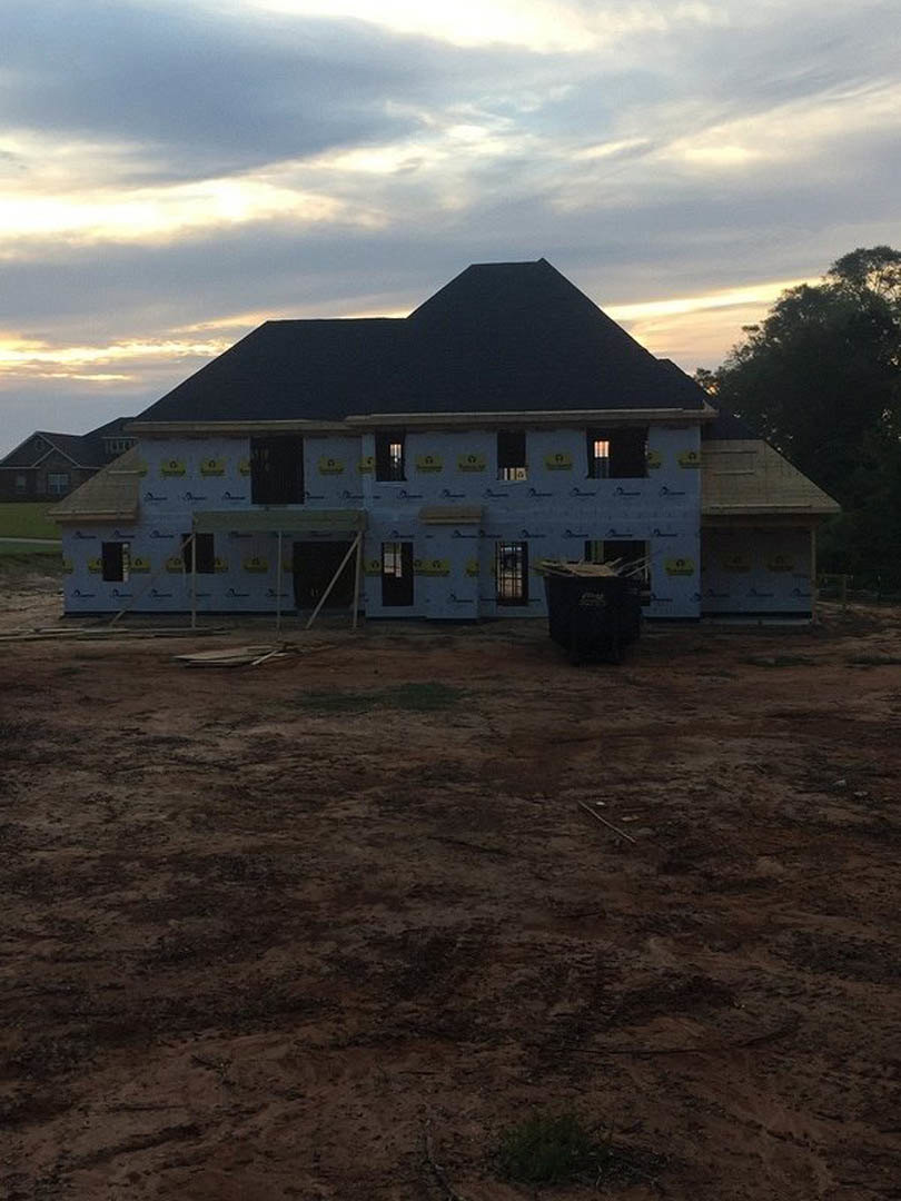 Partially built house with black roof, surrounded by dirt yard, scattered construction materials, black trash can, nearby tree, and mountain landscape under cloudy sky