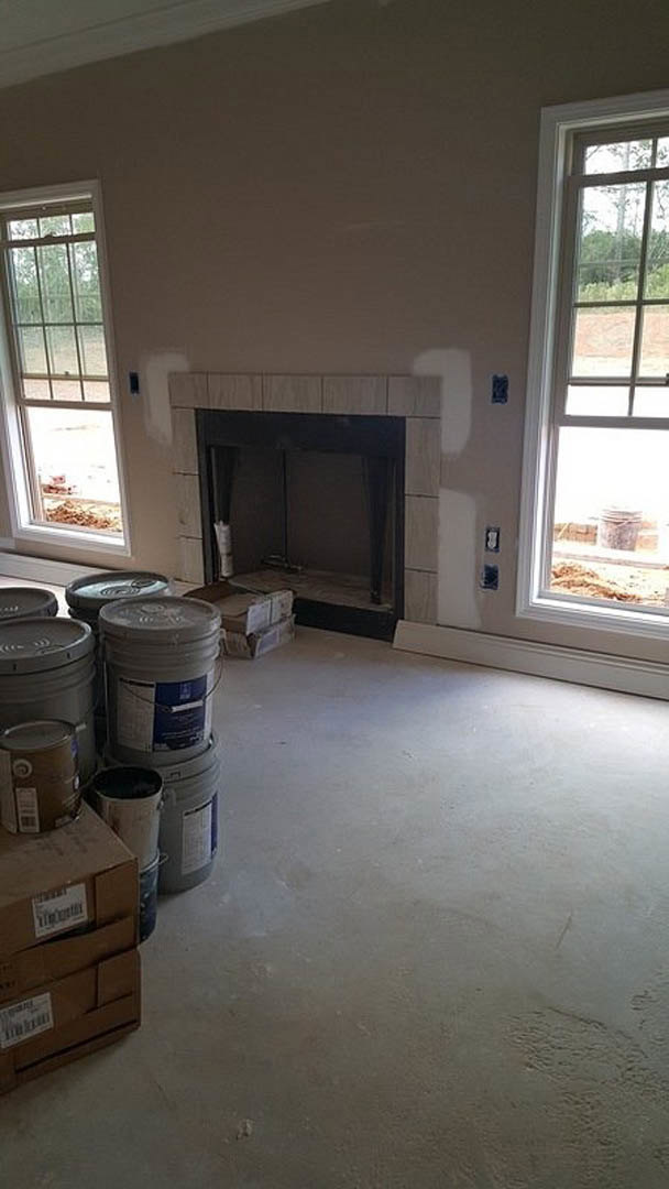 Living room with white-framed window, plaster walls, stone fireplace, hardwood flooring, and several paint buckets scattered near the hearth.