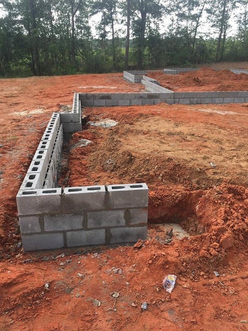 Excavated foundation with concrete block wall under construction, surrounded by dirt, grass, and trees