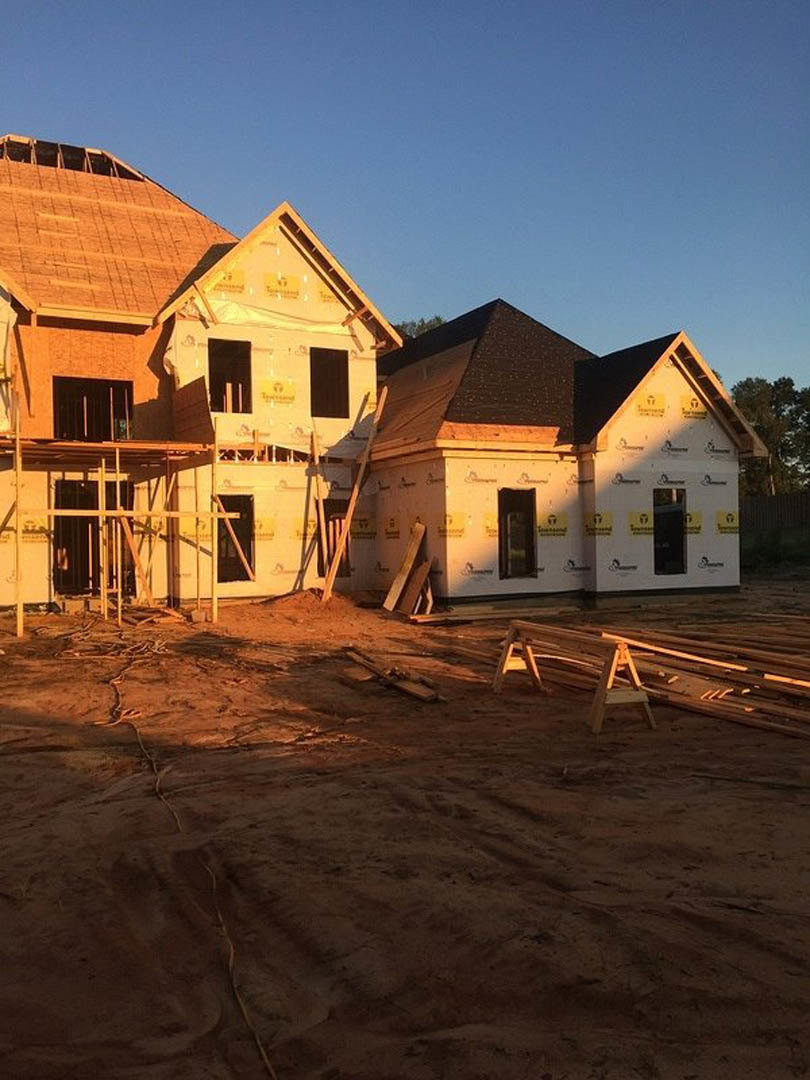 Framed house under construction with scaffolding, exposed wood beams, pile of lumber, small ladder, and neighboring homes under clear blue sky