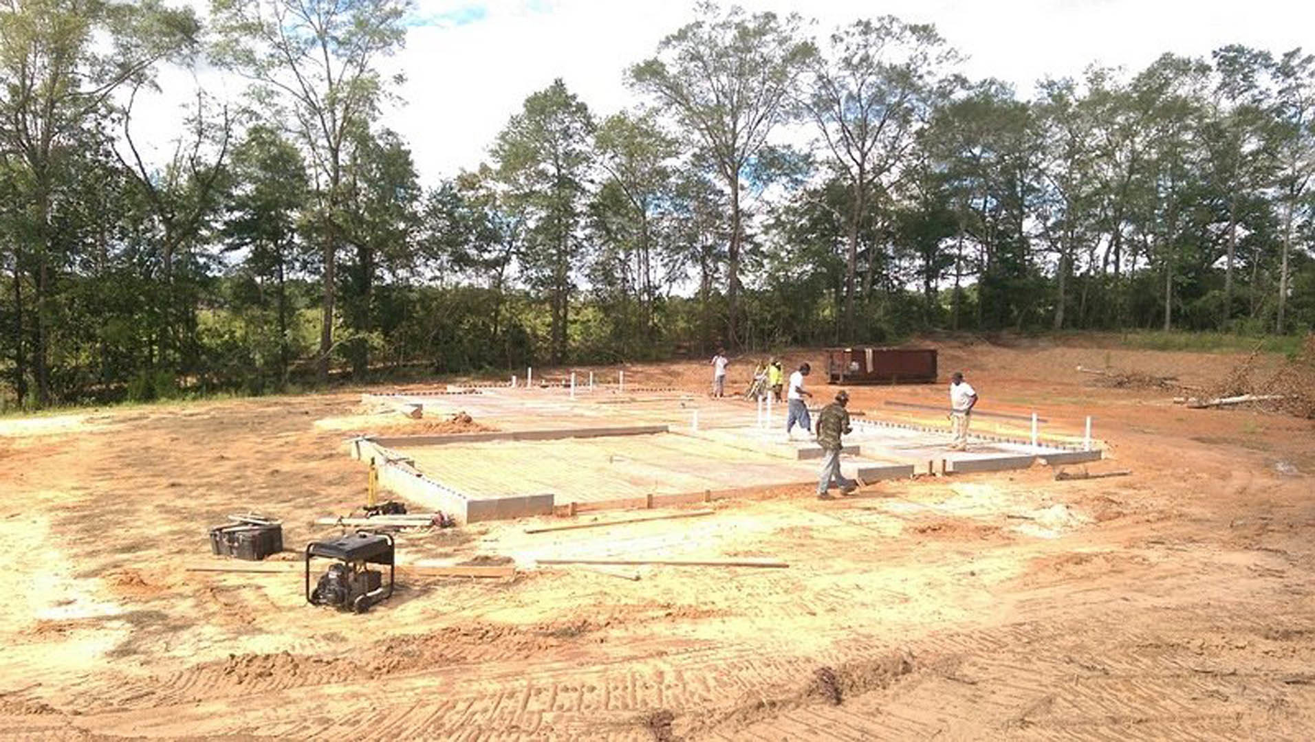 Group of people gathered on dirt ground near concrete foundation, surrounded by trees under open sky