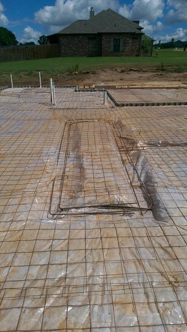 Metal-framed structure on a concrete slab foundation, partially covered with wire mesh and tarp, chimney visible on roof, wooden fence and trees in background under cloudy sky.