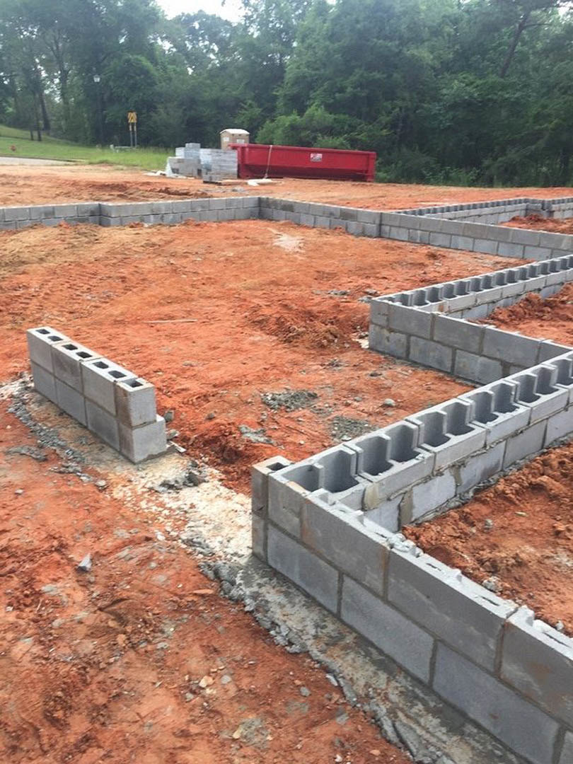 Concrete foundation corner surrounded by soil, stacked blocks, and a partially built brick wall; red dump truck and container with logo parked near trees.
