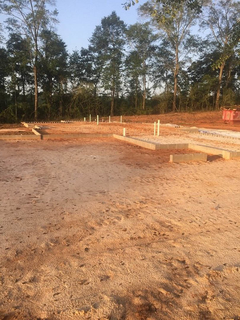 Dirt construction site with white poles, concrete block wall, and trees in the background