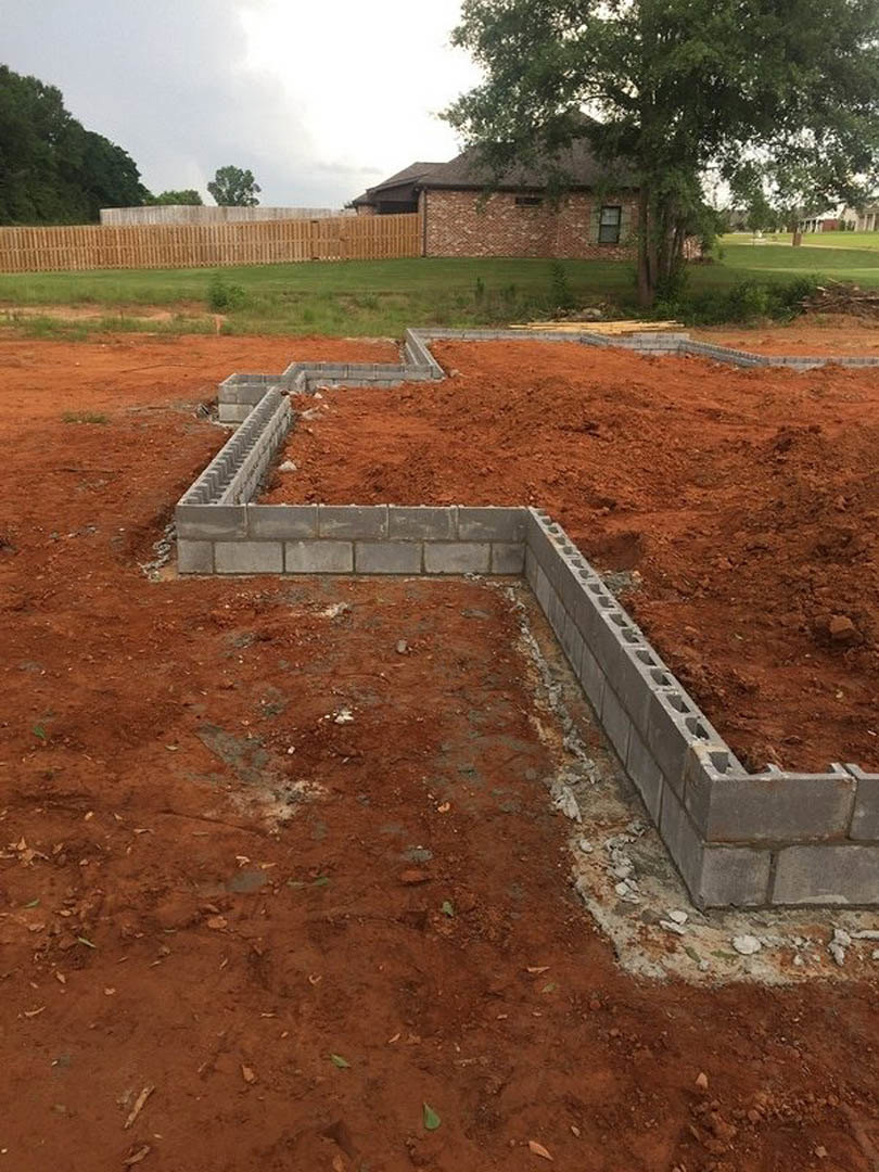 Concrete foundation with stacked blocks in a grassy yard, bordered by a wooden fence and mature trees, with a brick building and window visible in the background.