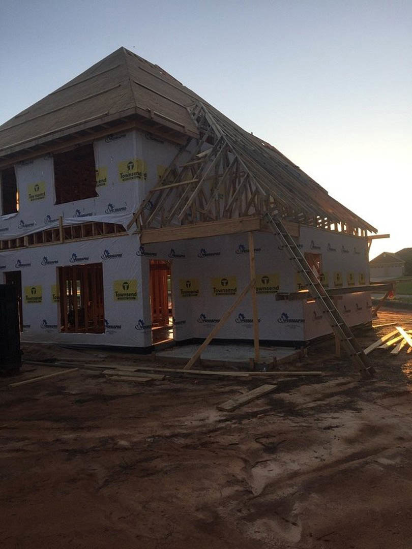 Dark brown brick exterior of a house under construction, dirt ground with scattered wood planks, ladder leaning near a window, close-up views of door and logo.