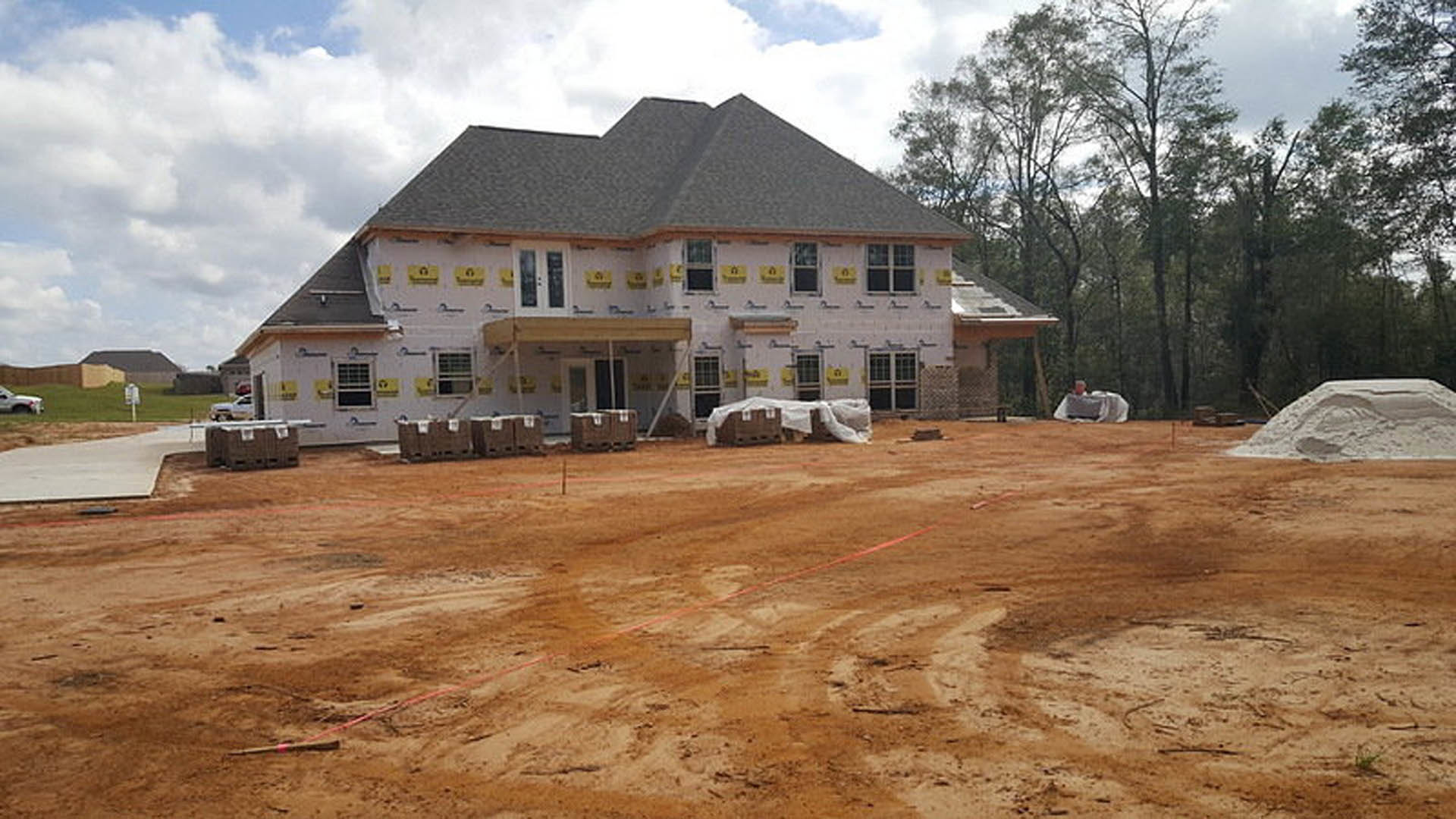 Two-story house under construction with exposed framing, piles of dirt in the foreground, yellow caution signs, plastic storage boxes, grid-patterned window, and trees in the
