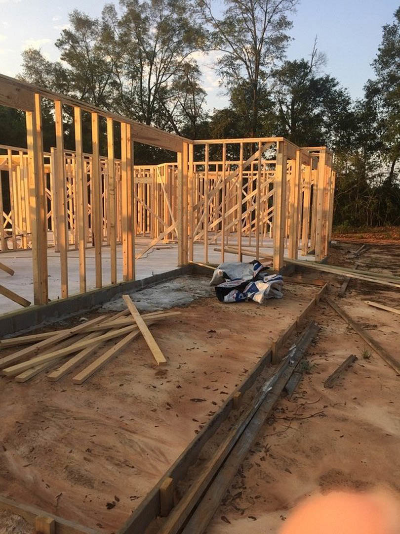 Wood-framed house under construction with exposed beams, piles of lumber on dirt ground, partially built exterior walls, and trees in the background