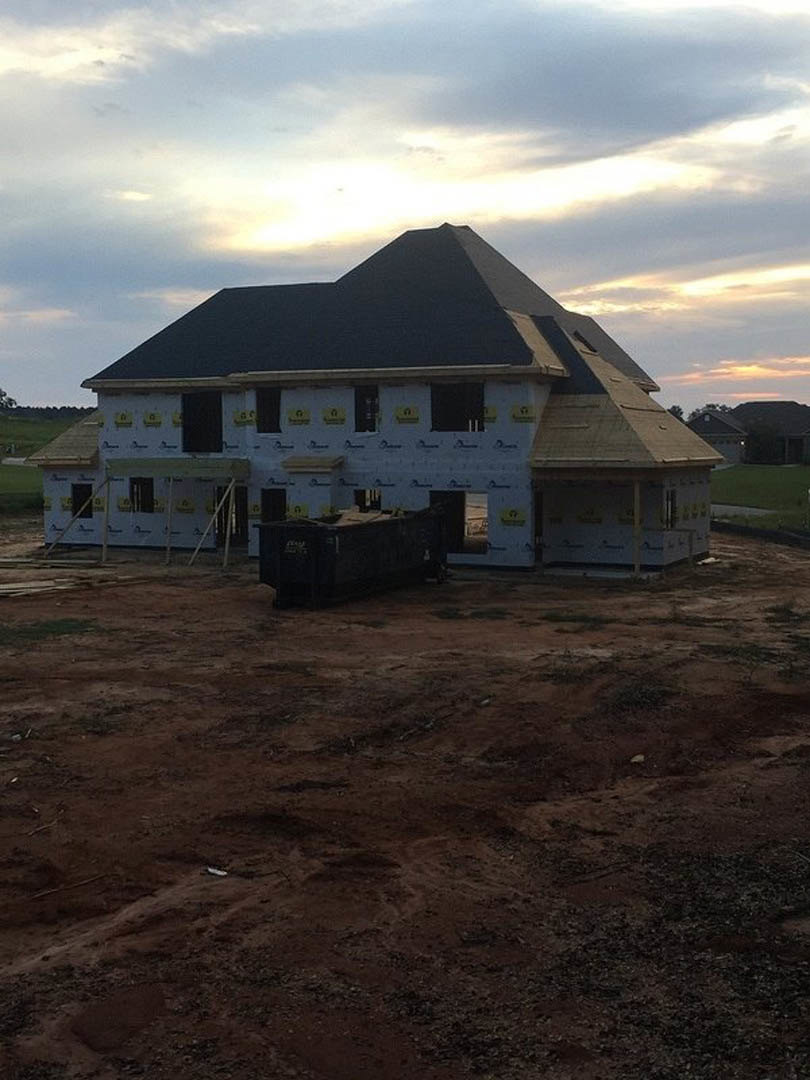 White custom home under construction with black roof, yellow construction signs on exterior wall, large dirt yard with scattered containers, grassy rural landscape under partly