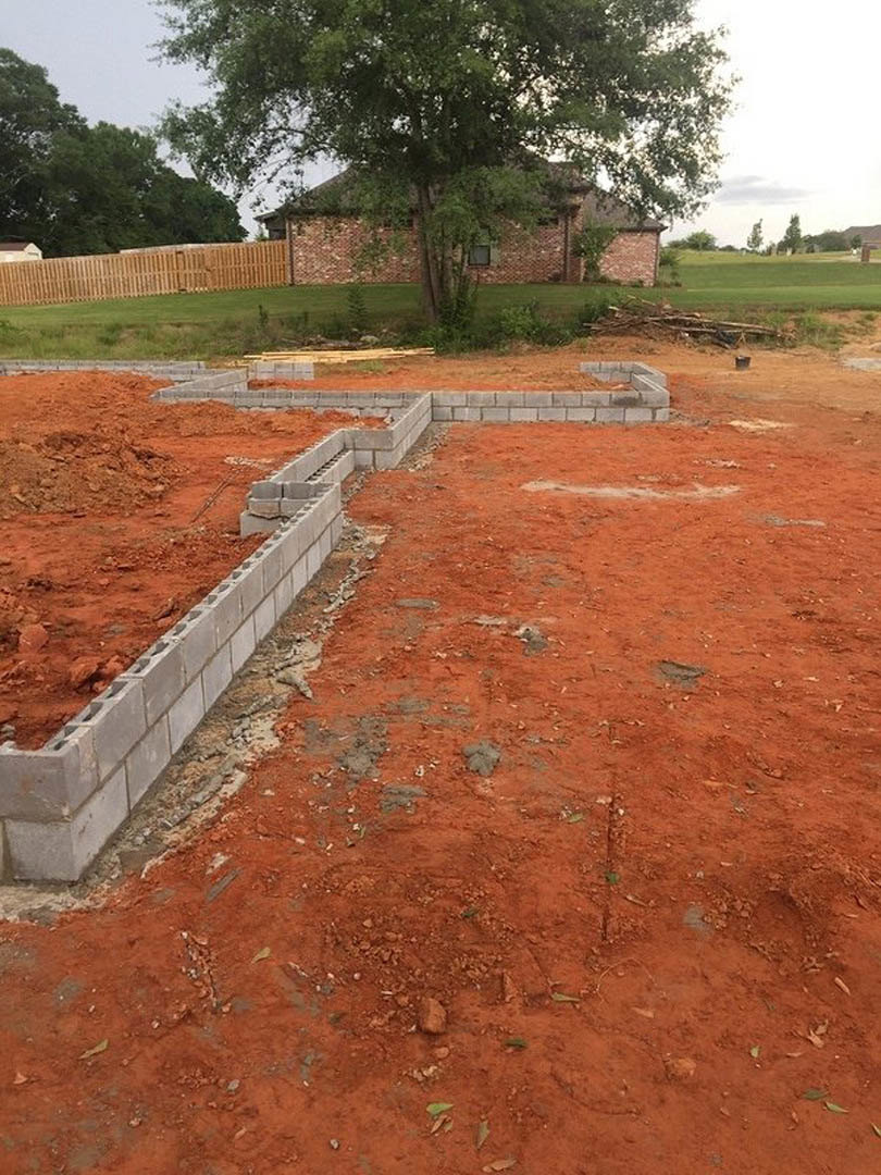 Brick house under construction with exposed foundation, grassy yard, wooden fence, and mature tree in front