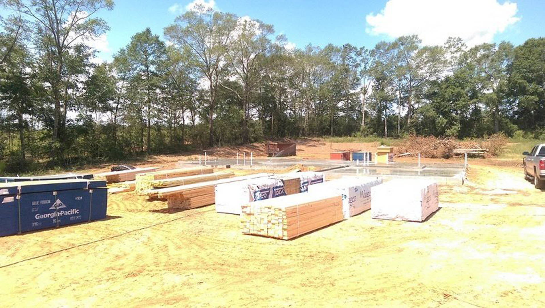 Framed wooden structure under construction, stacks of lumber on dirt ground, white building with fence in background, cloudy sky overhead.