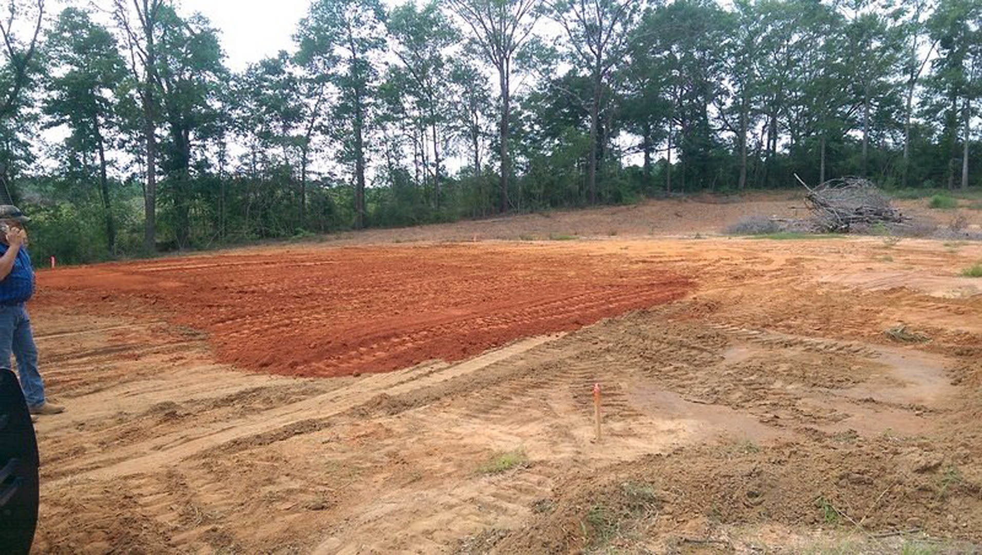 Dirt field bordered by trees under open sky, with a dirt path and a person in blue jeans walking along the edge