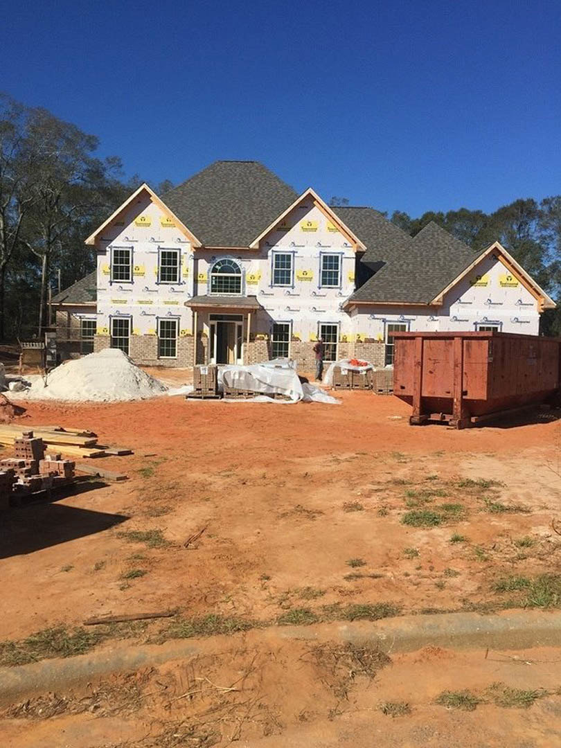 Partially built house with exposed framing, large wooden container on a pallet, construction materials scattered on the ground, snow-covered car parked nearby, tree in the