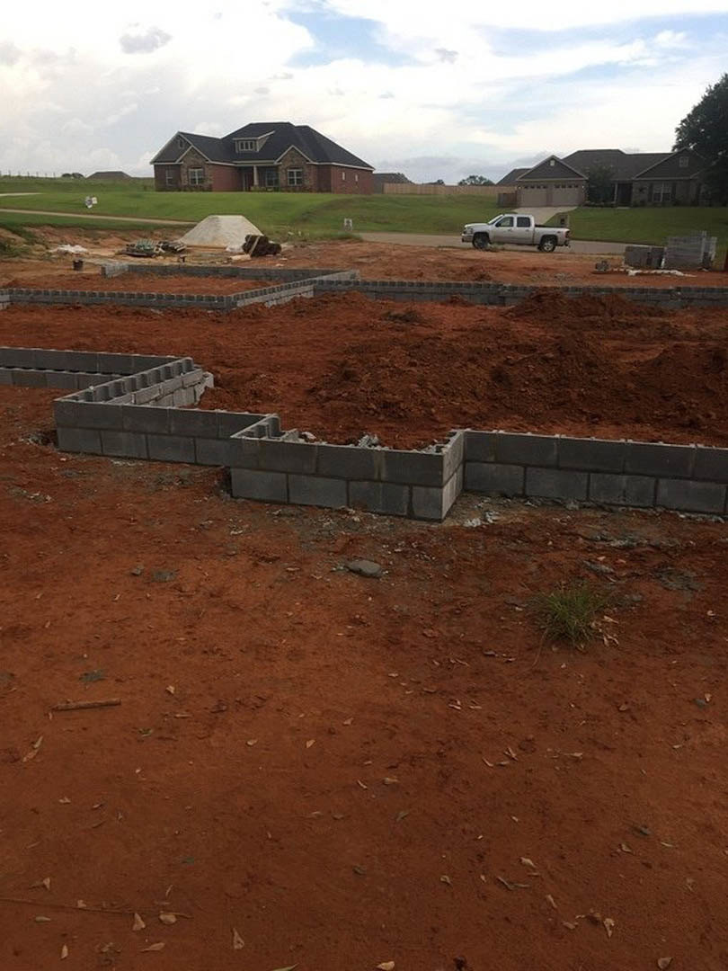 Partially built house with black roof and brick walls, white truck parked on dirt lot, large white rock in foreground, cloudy sky overhead, scattered trees and mountains in