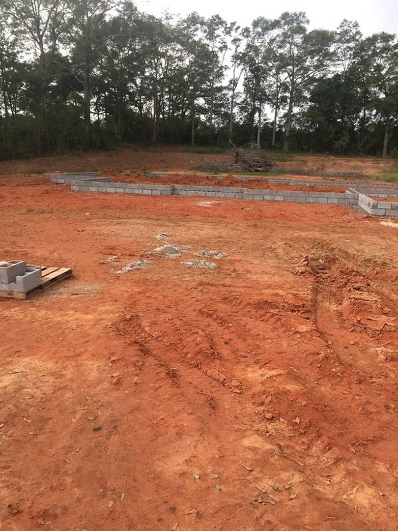 Bricks stacked on pallets beside a partially built brick wall, dirt ground, scattered construction materials, and trees in the background