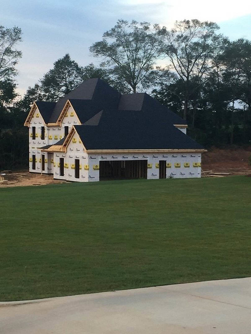 Partially built house with exposed framing and unfinished exterior, surrounded by green lawn and mature trees under a cloudy sky