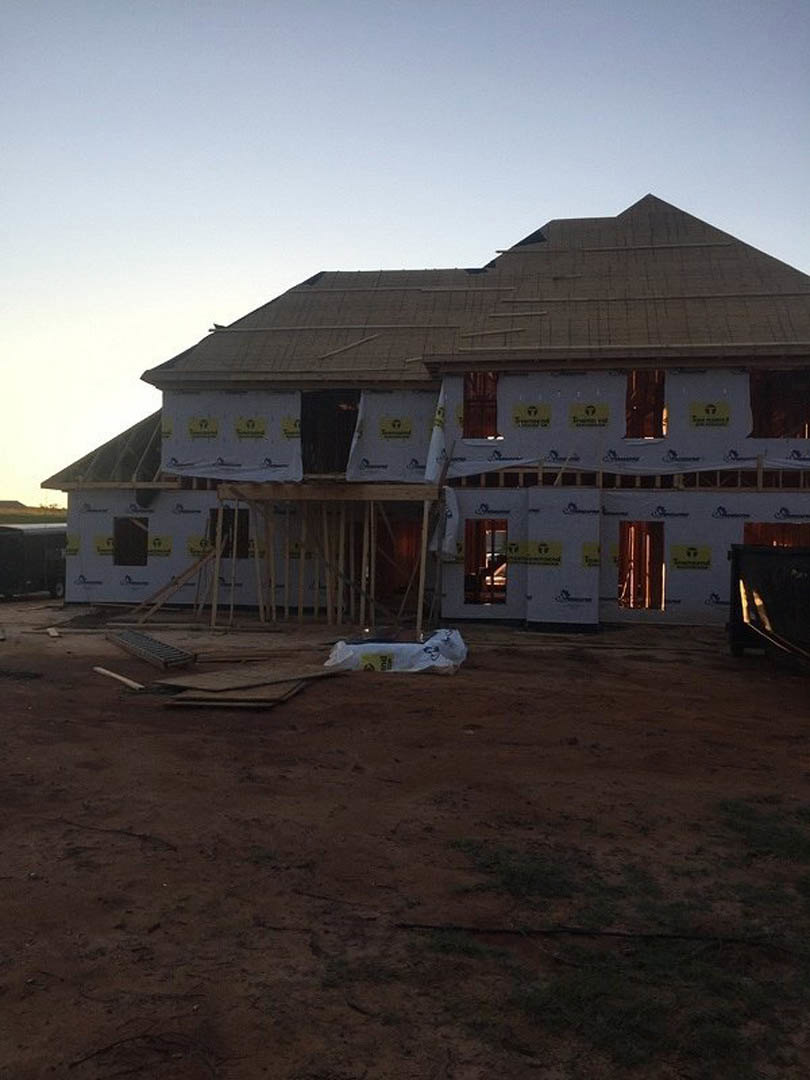 Partially built house with exposed framing, several installed windows, scaffolding along exterior walls, dirt yard with construction materials, under clear sky