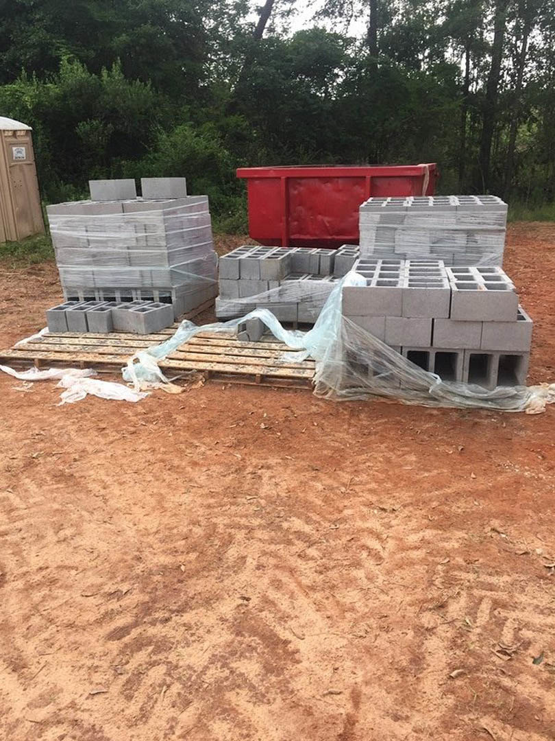 Grey bricks stacked on wooden pallets beside a red dumpster and white bricks, concrete blocks wrapped in plastic, brown box with white label, dirt ground, netting, and scattered