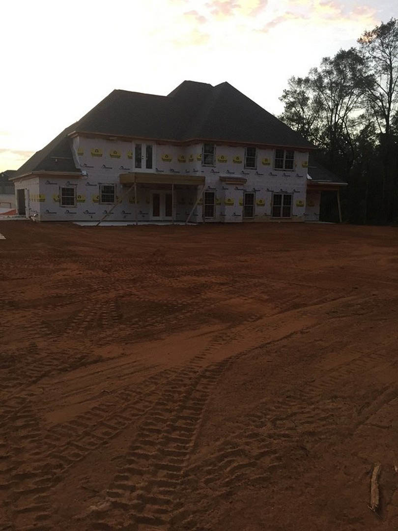 Framed house under construction surrounded by dirt and tire tracks, yellow signs attached to exterior walls, trees in background, cloudy sky overhead