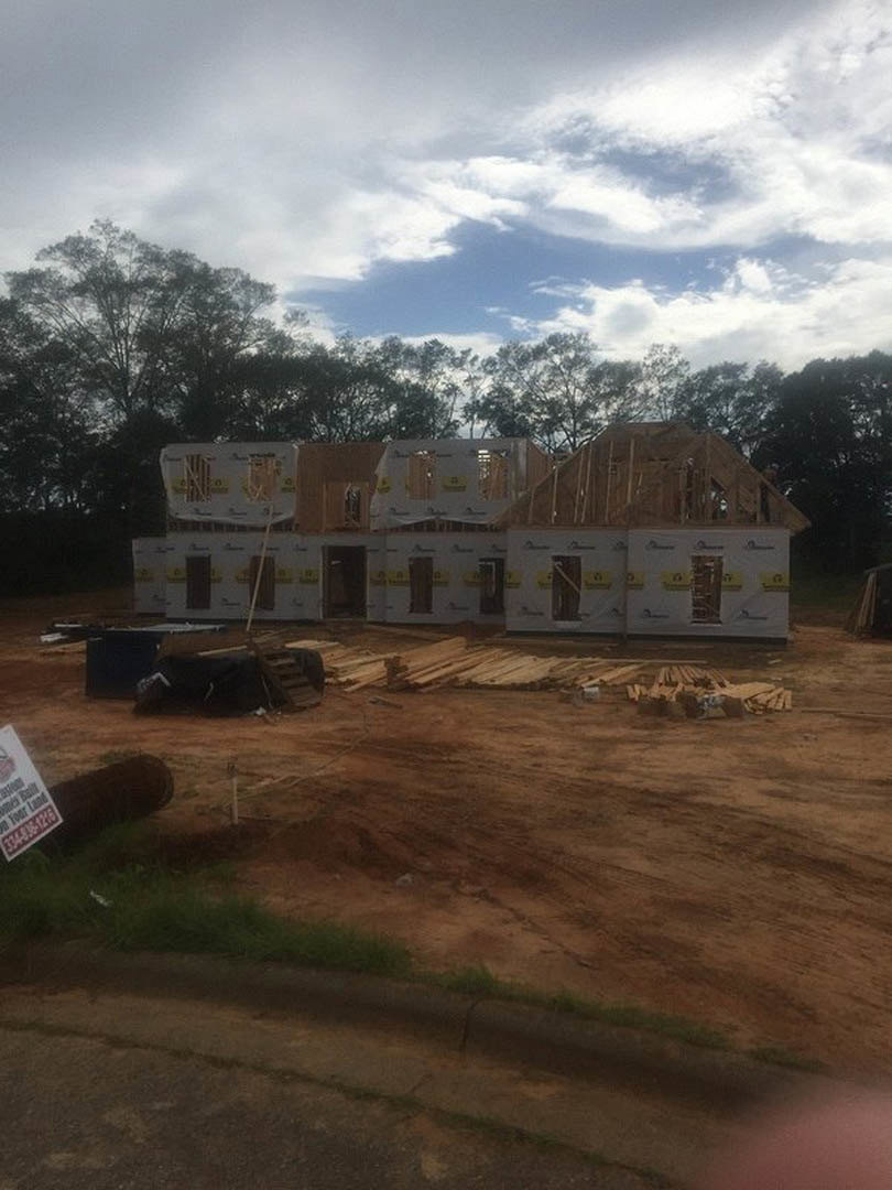 Framed wooden house under construction on grassy lot, stacks of lumber in foreground, trees and blue sky with scattered clouds in background